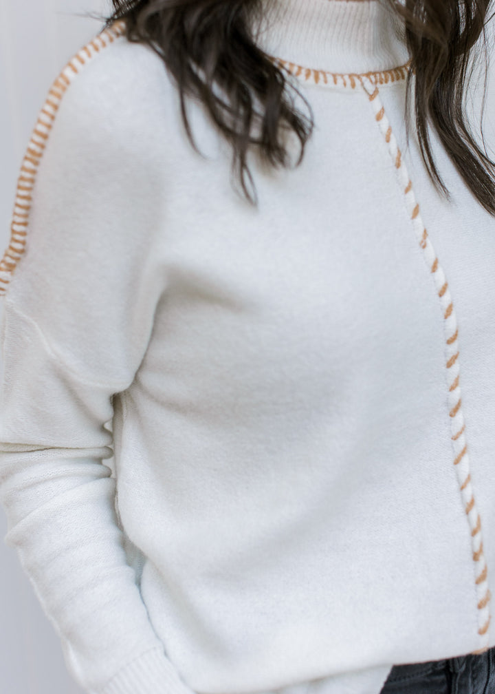 Close up of camel stitch work on exposed hem of a cream sweater with long sleeves and a mock neck. 