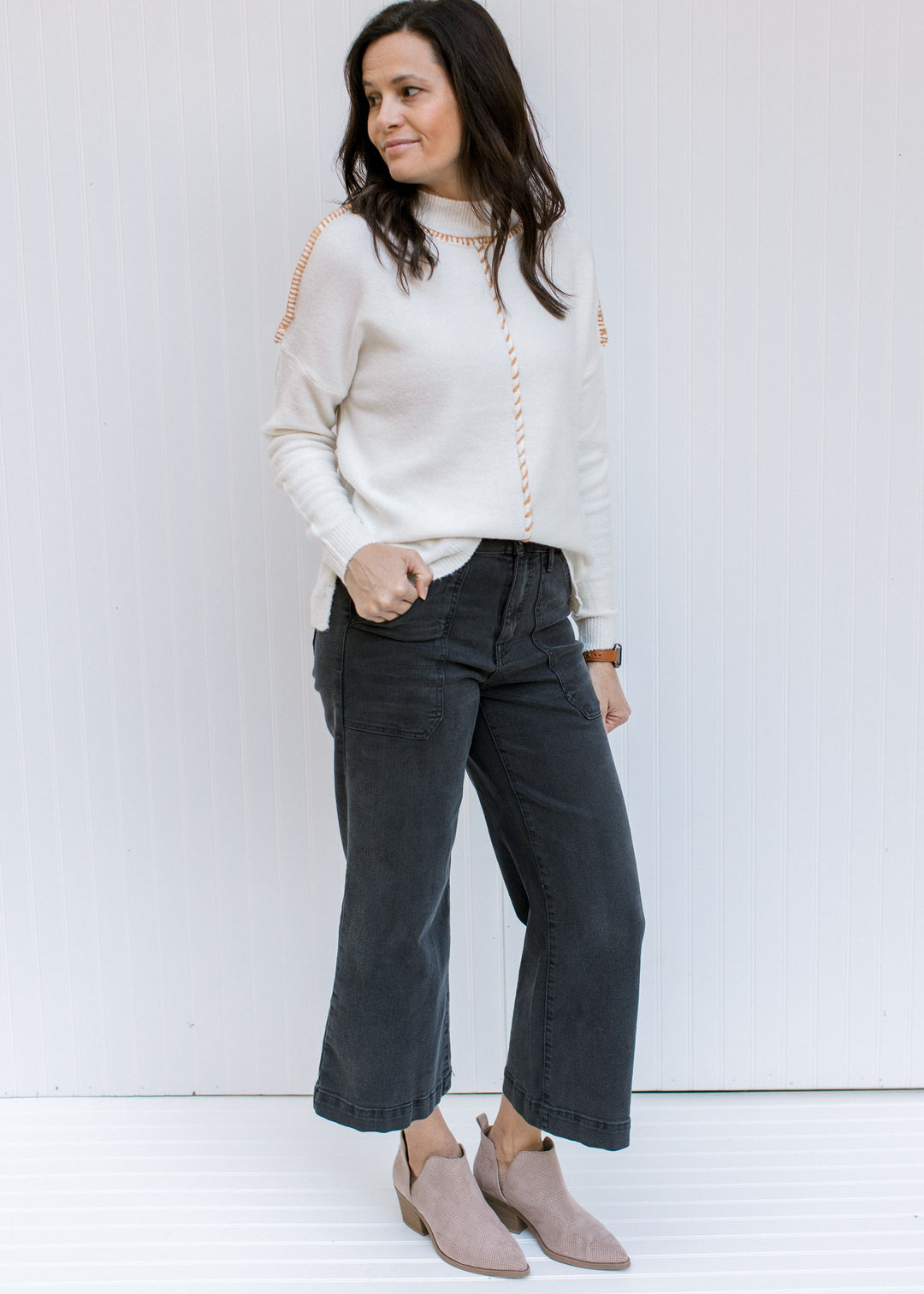 Model wearing jeans with a cream sweater with a camel stitch work on exposed hem and long sleeves. 