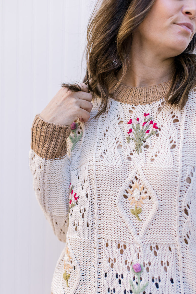 Close up of colorful stitched flowers on a loose knit cream sweater with long sleeves.