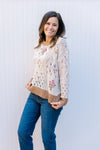 Model wearing jeans and a cream sweater with stitched colorful flowers, long sleeves and brown hem.