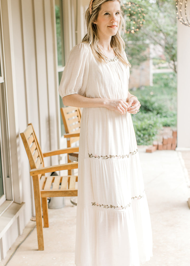 Model wearing a cream maxi with green and pink floral embroidery at neck, waist and tiers.