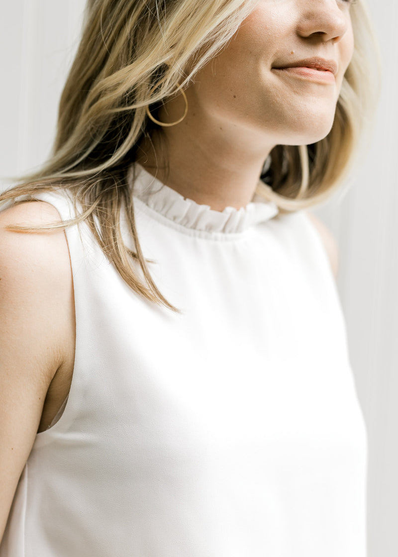 Close up view of ruffle at the neck of a sleeveless ivory top. 