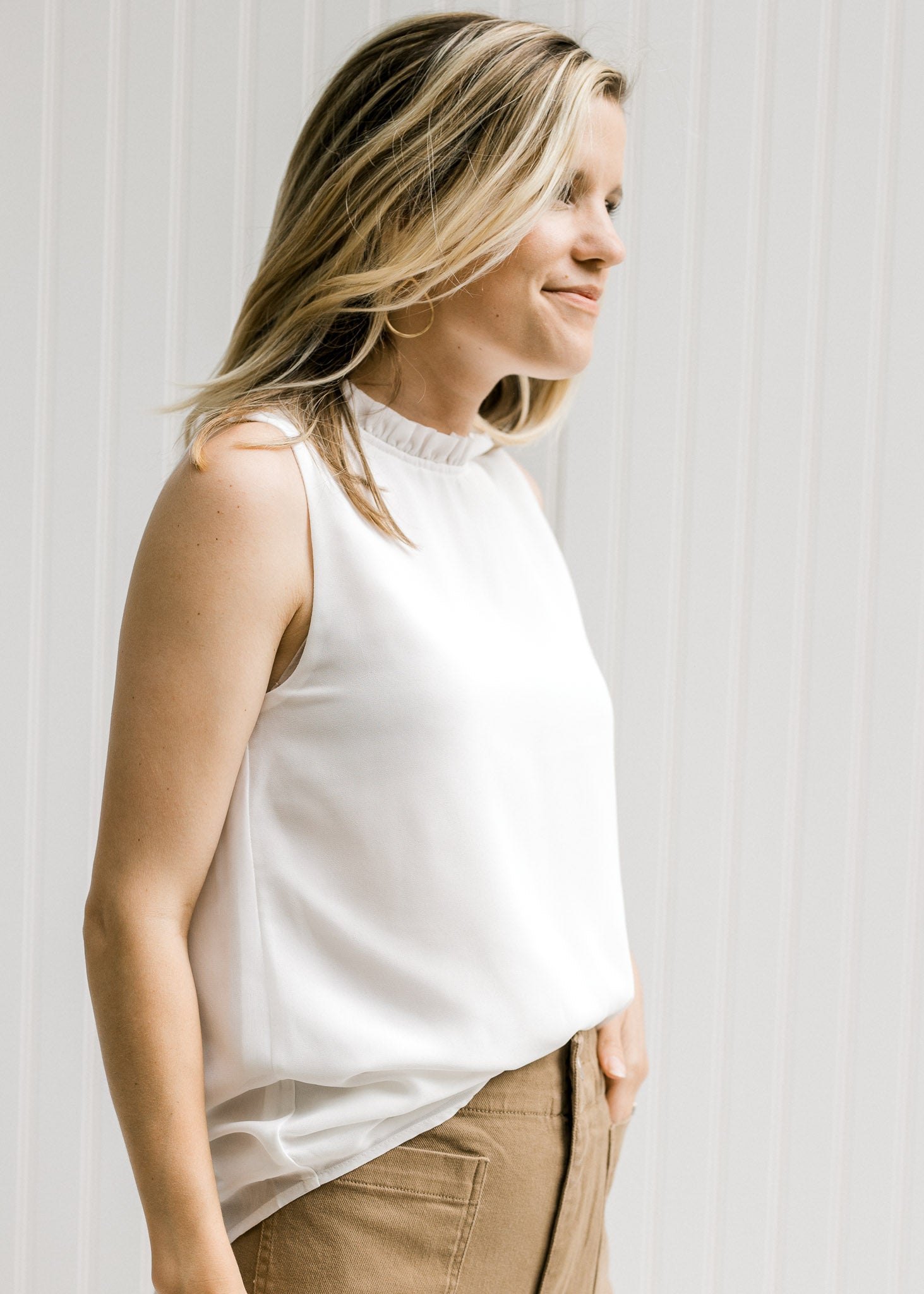 Model wearing an ivory sleeveless top with a ruffle at the neck.