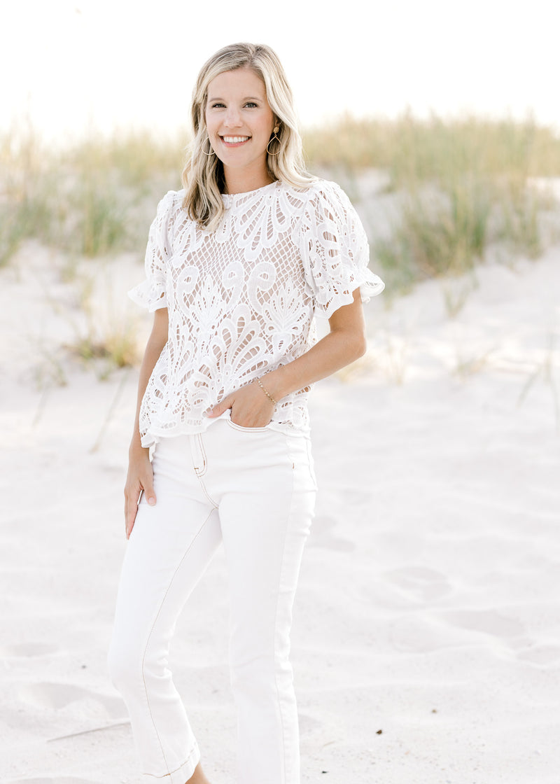 Model wearing white jeans with a nude short sleeve top with an ivory lace overlay and a round neck. 