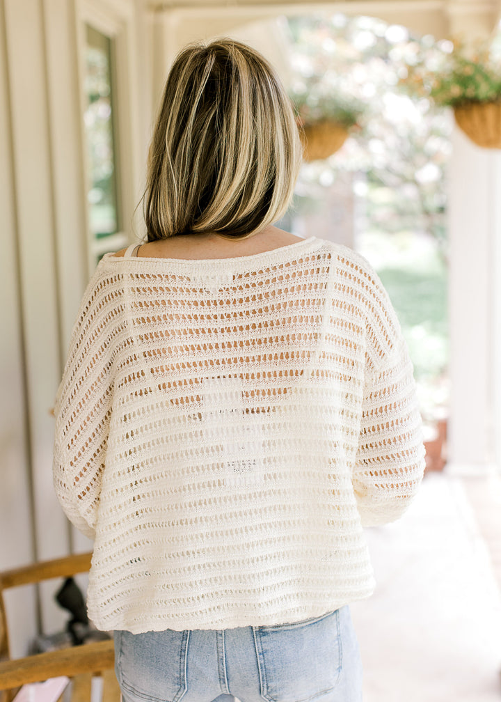 Back view of Model in an ivory open knit blouse with a weave design, long sleeves and boat neck.