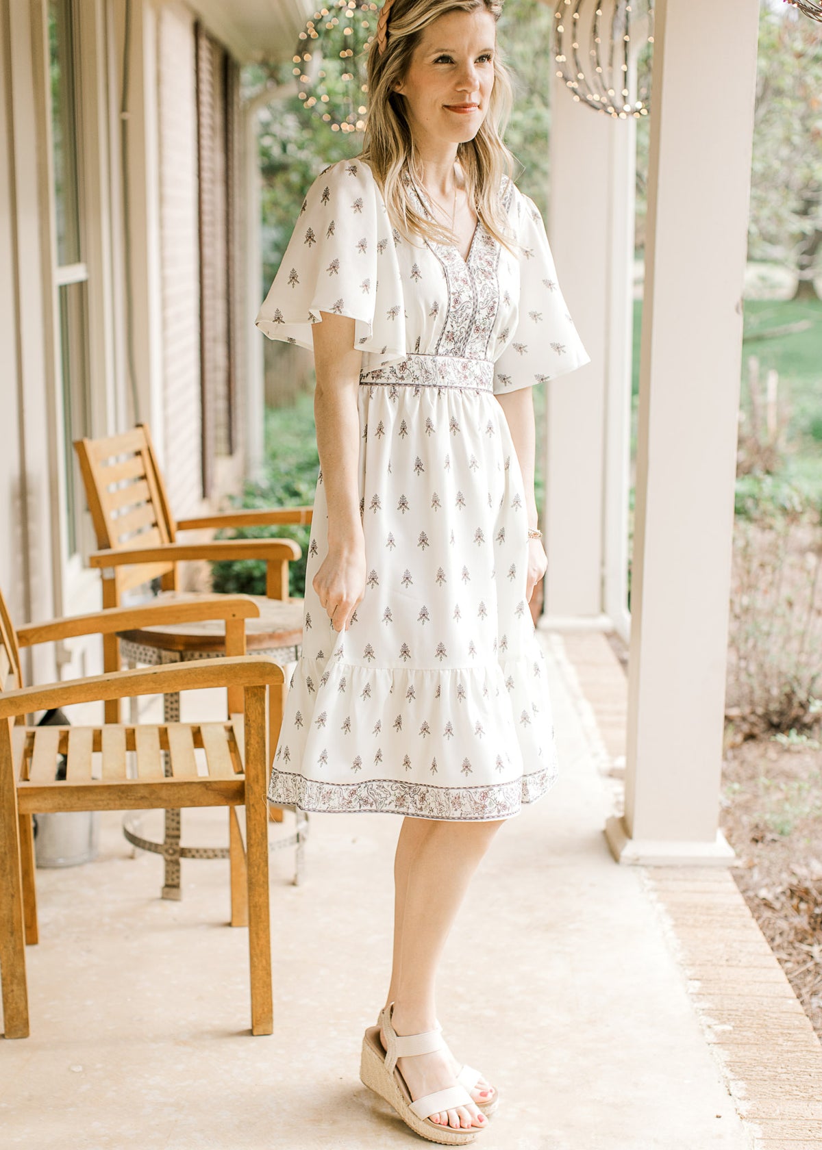 Model wearing wedges and a pale cream dress with mauve and green floral, short sleeves and v-neck.