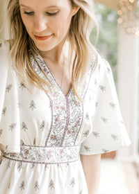 Close up of detail trim at  v-neck bodice, waist, and hem of a short sleeve ivory dress. 