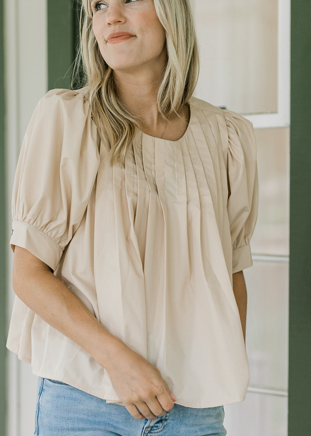 Model wearing a short sleeve ivory top with a round neck and pleating at neck and shoulder. 