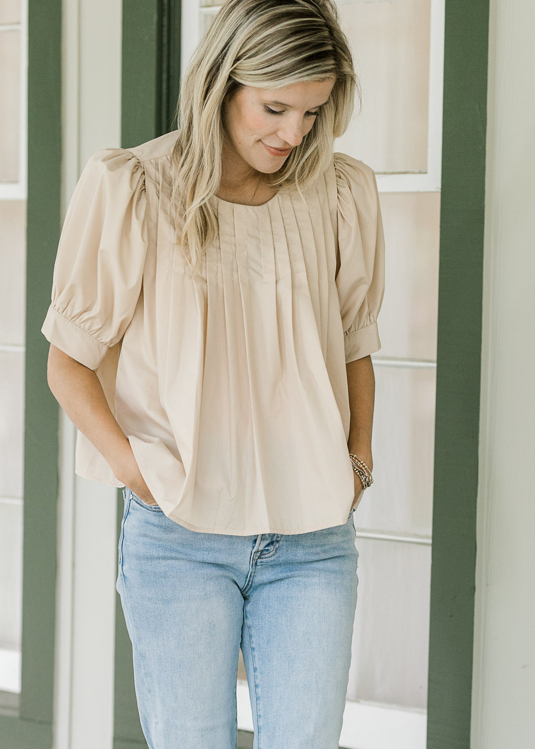 Model wearing jeans and an ivory top with a round neck and pleating at neck and shoulder. 
