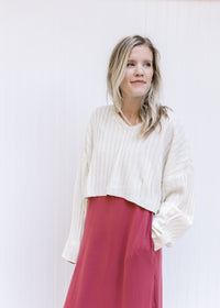 Model wearing a ribbed ivory sweater with a wide v-neck, a cropped fit and long sleeves with a maxi.