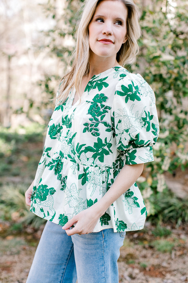 Model wearing a white short sleeve top with green foliage, v-neck and a ruffled peplum fit.