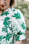 Close up of short sleeve with pleated shoulder and cuff on a white top with green foliage. 