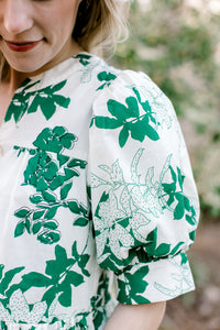 Close up of short sleeve with pleated shoulder and cuff on a white top with green foliage. 