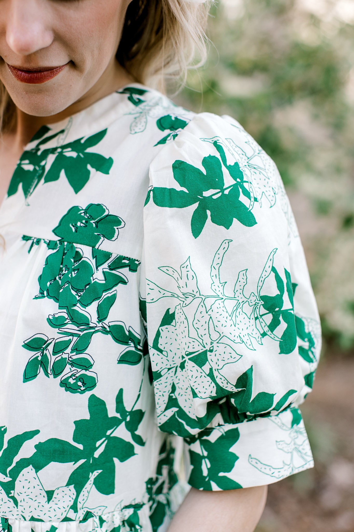 Close up of short sleeve with pleated shoulder and cuff on a white top with green foliage. 