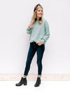 Model wearing jeans, with black booties and a jade top with long sleeves and rolled hem detail. 