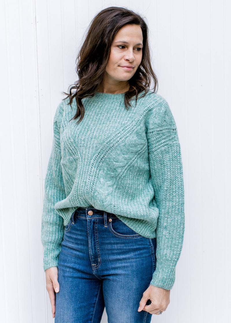 Model wearing a jade sweater with cable knit fabric, long sleeves and a round neck tucked in jeans.