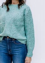 Close up of cable knit design on a jade sweater with long sleeves and a round neckline. 