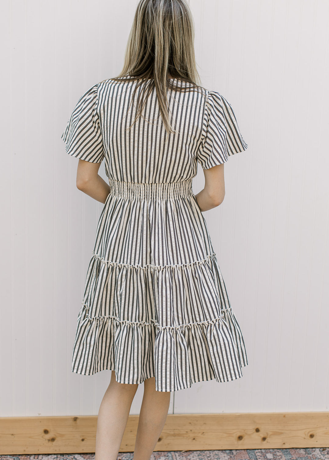 Back view of Model wearing an off-white dress with charcoal stripes, v-neck and short sleeves.