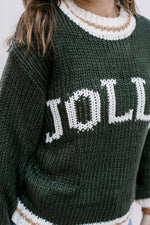 Close up of “JOLLY” written in cream across a hunter green knit sweater with long sleeves. 