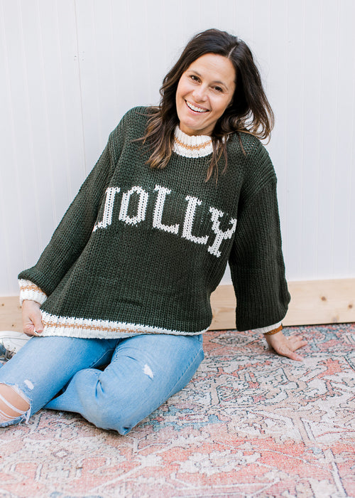 Model wearing a hunter green long sleeve sweater with “JOLLY” across the front in cream font. 