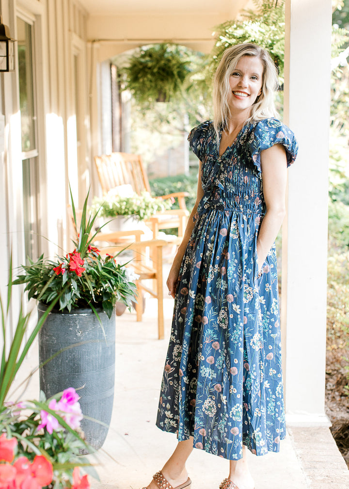 Model wearing wedges and a blue midi with bright floral, smocked bodice and short sleeves. 