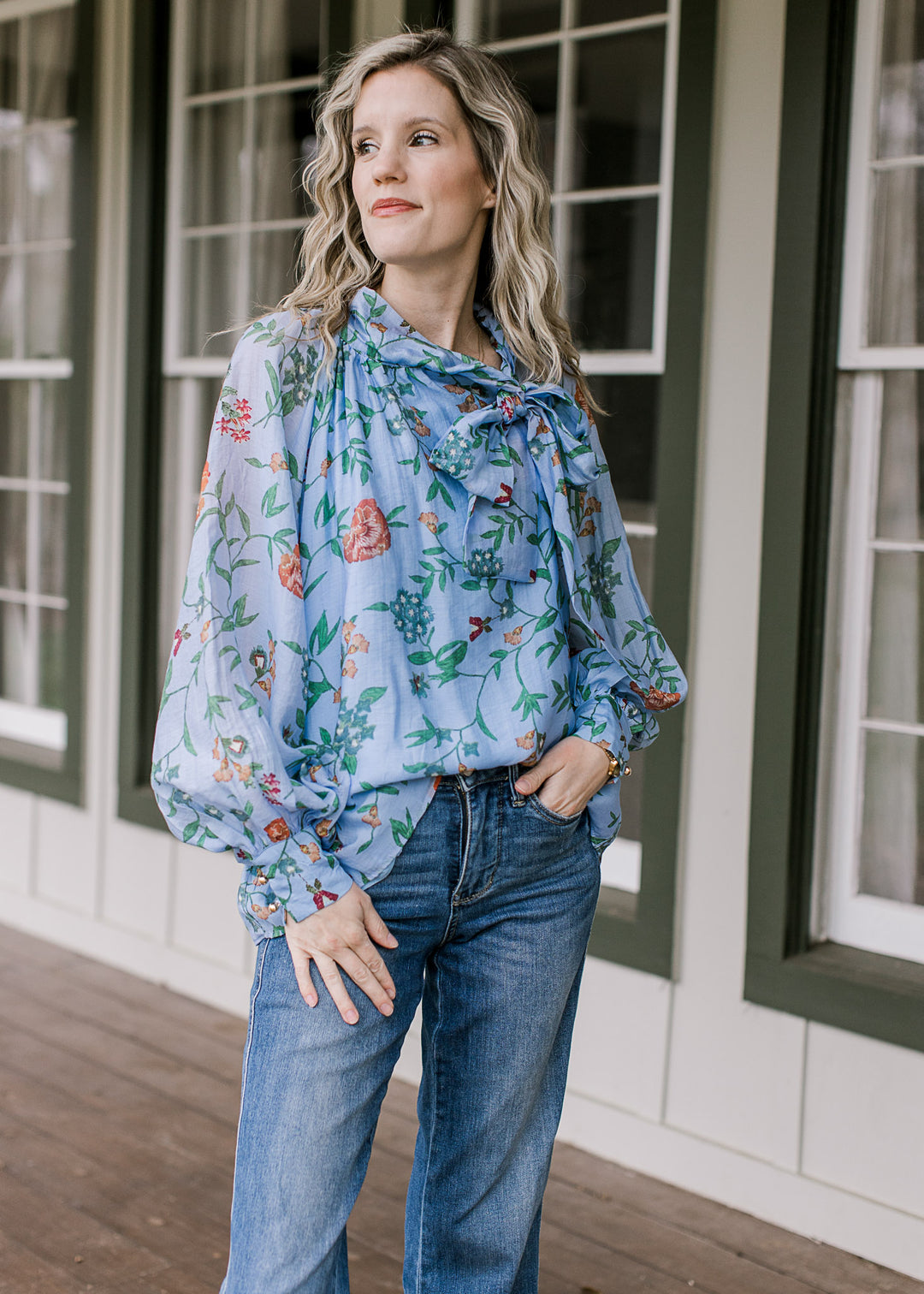 Model wearing jeans and a blue top with green and salmon floral, long sleeves and side tie at neck.