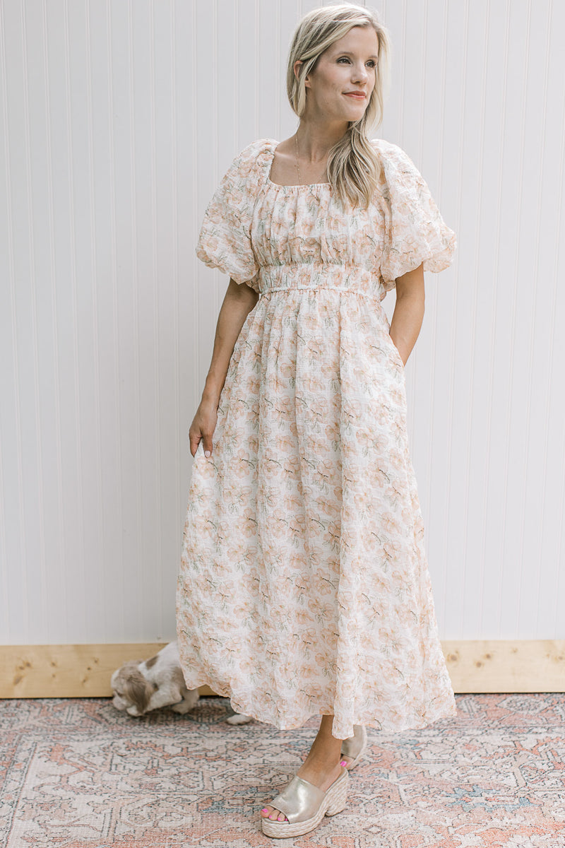 Model wearing a cream dress with peach and green flowers, bubble short sleeves and smocked waist.