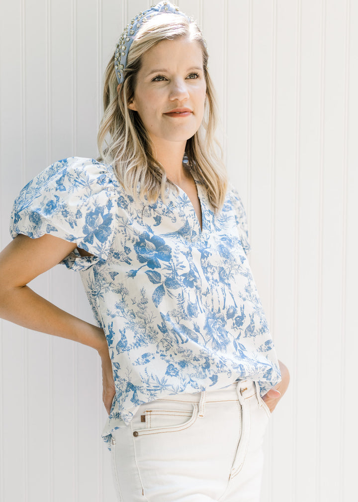 Model wearing a white v-neck top with blue floral and animal patterns and short puff sleeves. 