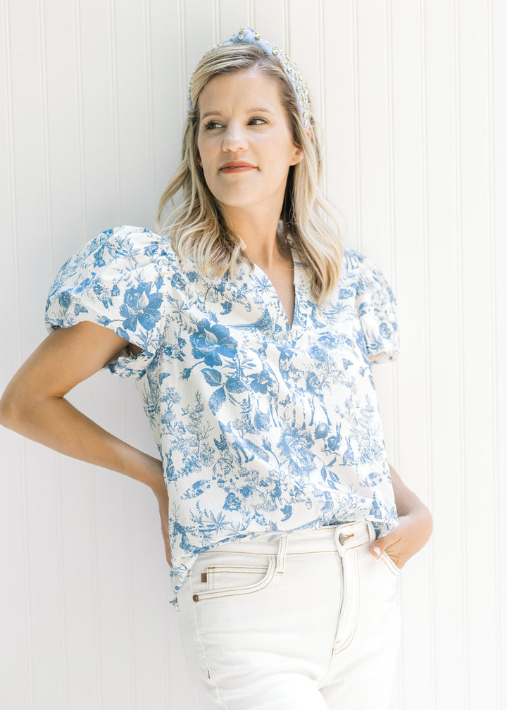 Model wearing white jeans with a white v-neck top with blue floral and animal patterns. 