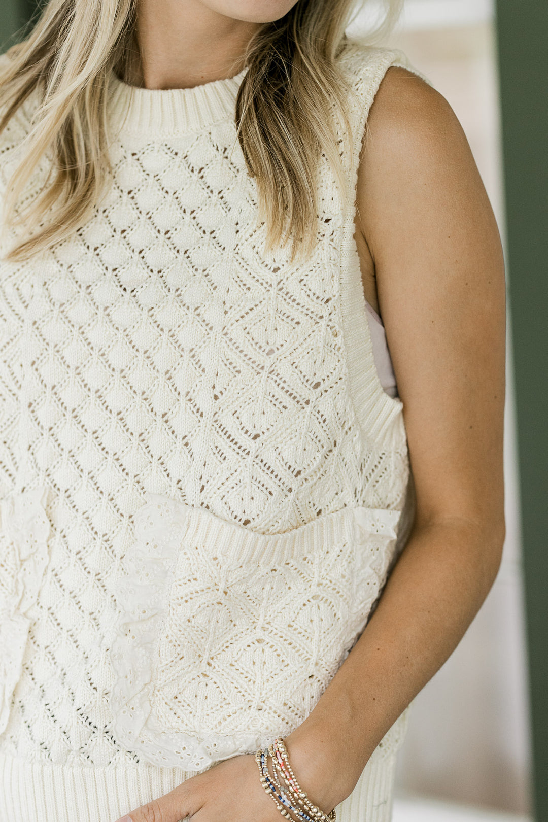 Close up of patch pockets lined with lace on a model in a cream knit sleeveless sweater. 