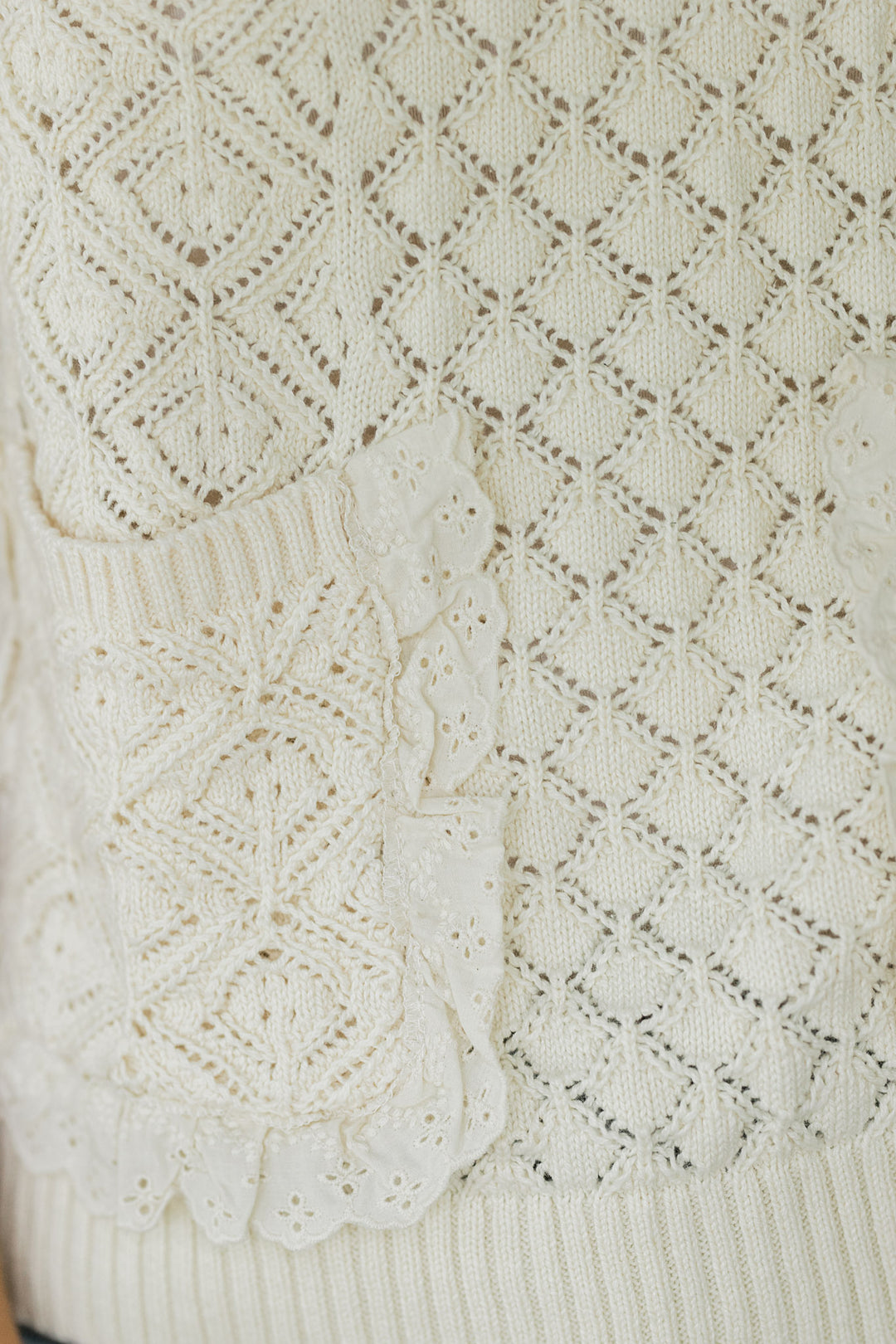 Close up of lace detail around the patch pocket of a cream knit sleeveless sweater. 