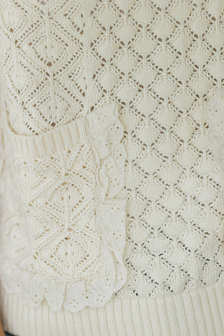 Close up of lace detail around the patch pocket of a cream knit sleeveless sweater. 