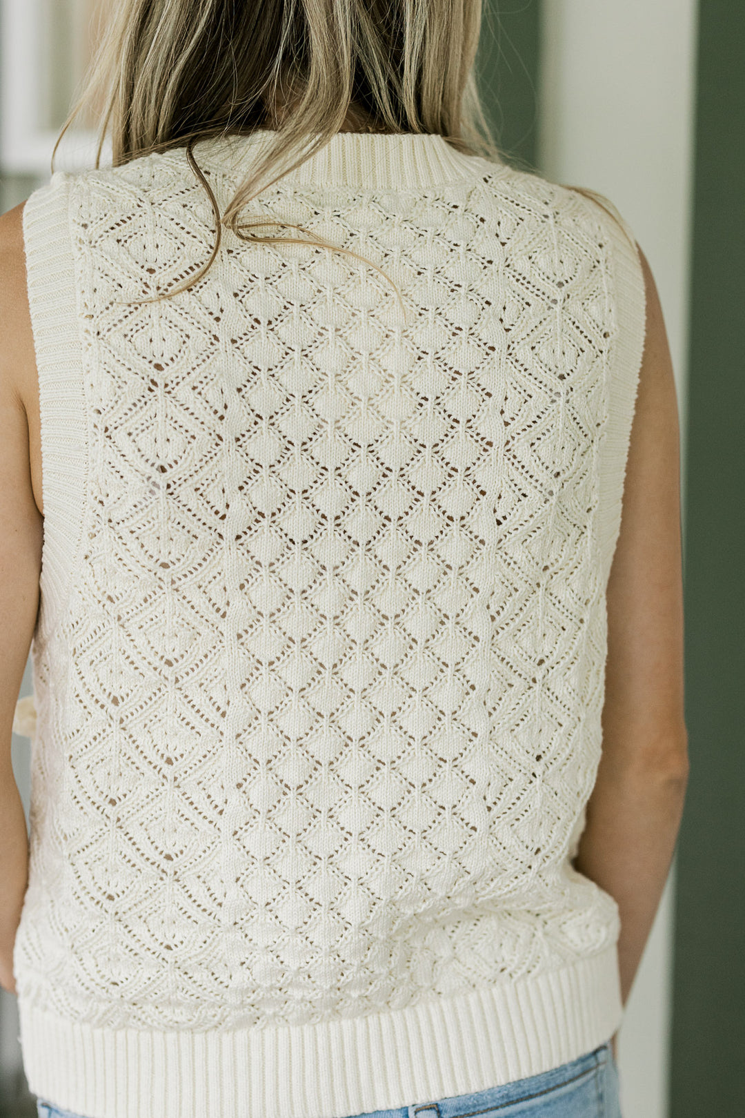 Back view of Model wearing a sleeveless cream sweater with ribbed detail at neck, sleeve and hem. 