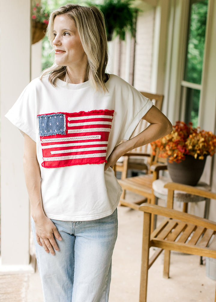Model wearing jeans and an off-white top with American flag graphic, round neck and  cap sleeves.