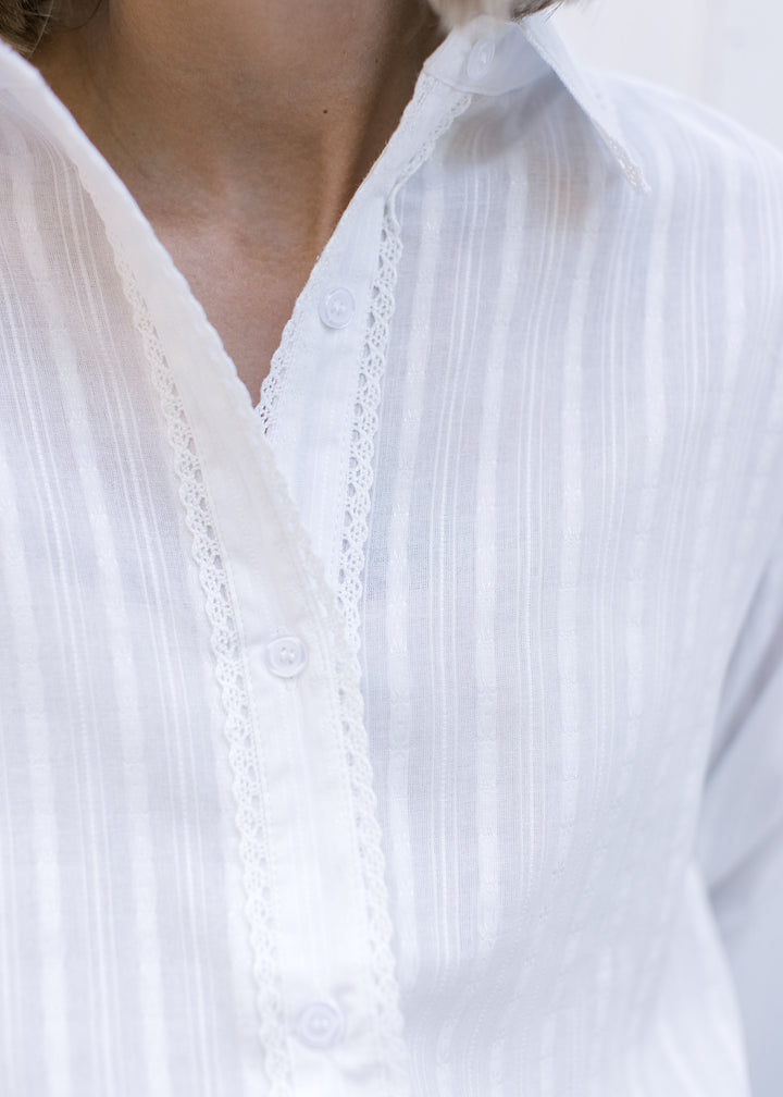 Close up of lace detail at button placket and subtle stripes on a white long sleeve top. 