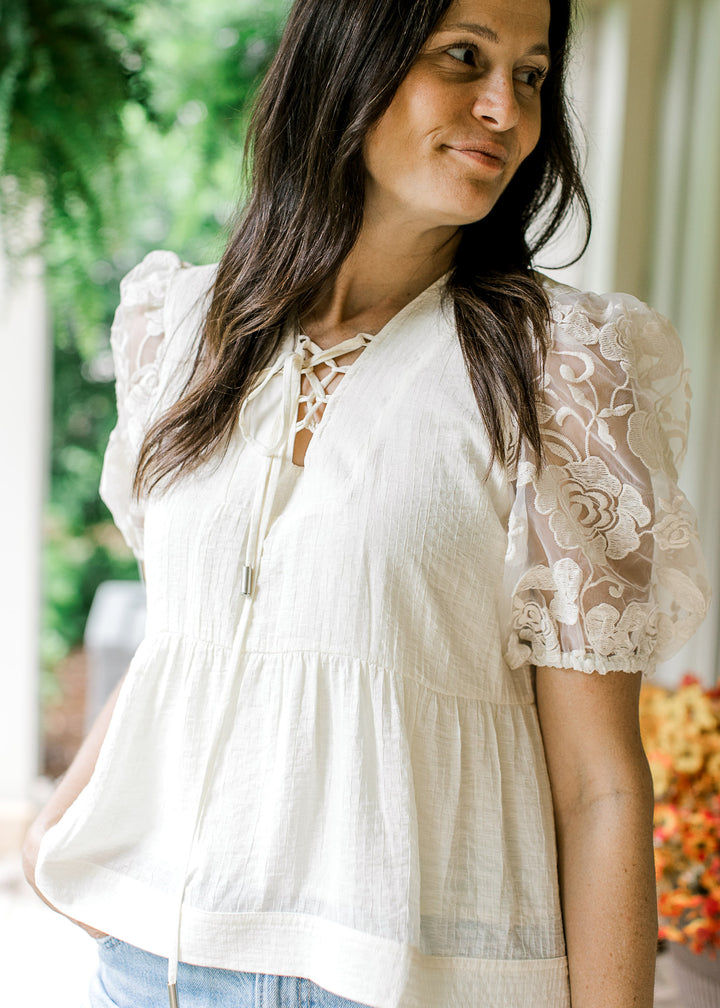 Model wearing a cream top with textured bodice, lace-up V-neck and sheer floral short sleeves.