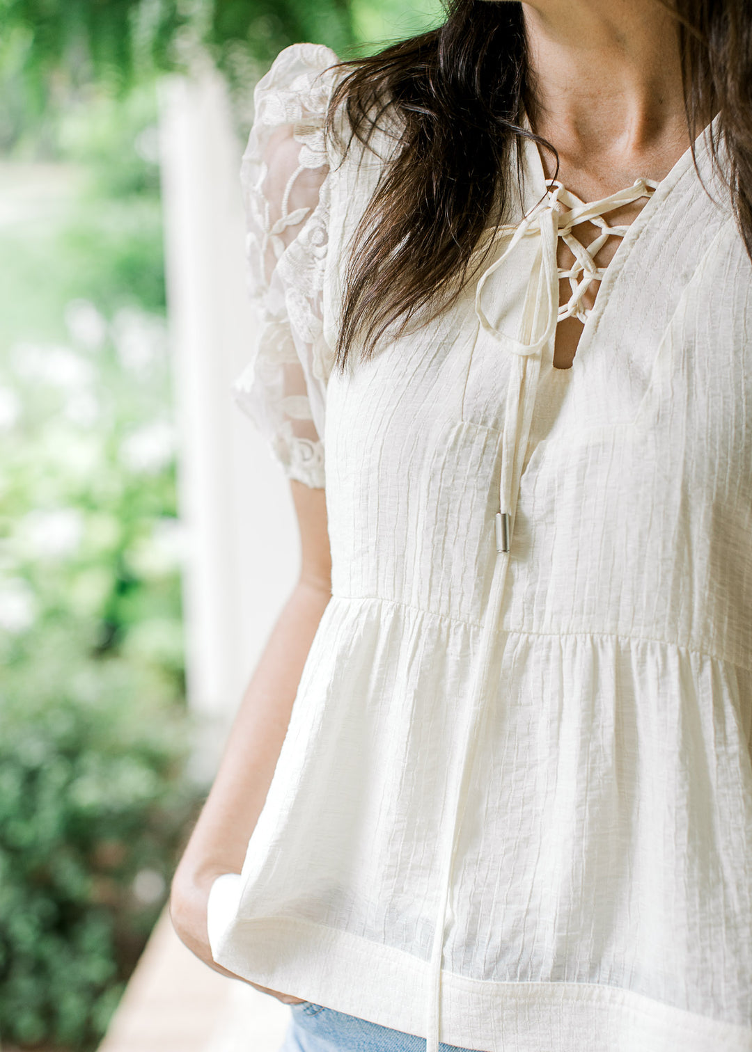 Model wearing a cream babydoll top with texturing, lace-up V-neck and sheer floral short sleeves.