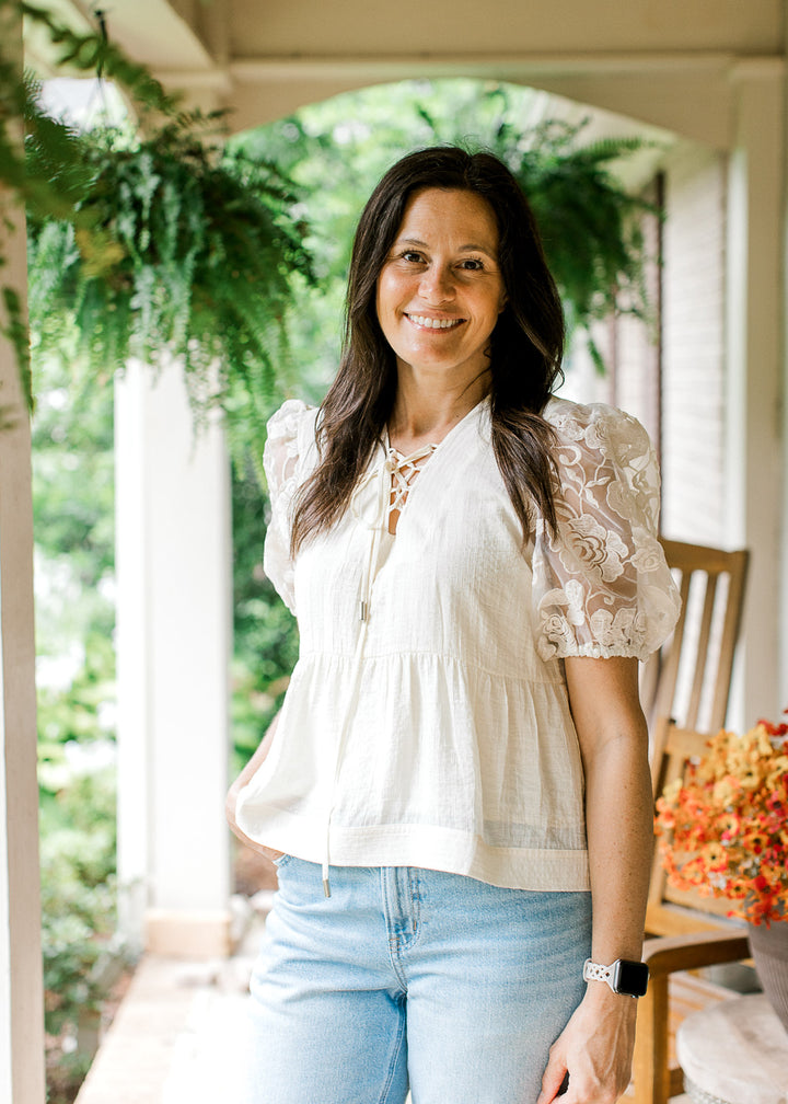 Model wearing jeans and a cream top with textured bodice, lace-up V-neck and floral short sleeves.