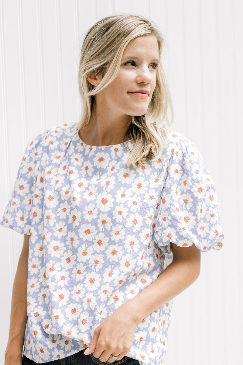 Model wearing a lavender top with a cream daisy pattern, round neck and short puff sleeves. 