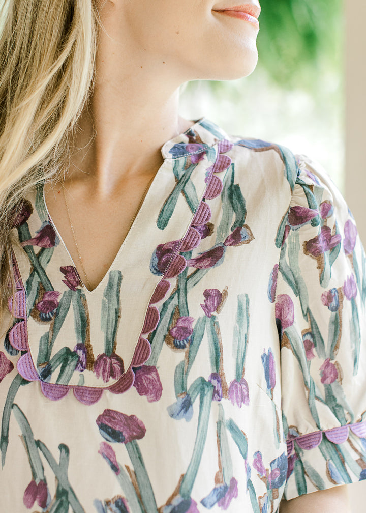 Close up of lavender scalloped lace on bib collar and cuff of a short sleeve cream top with floral. 