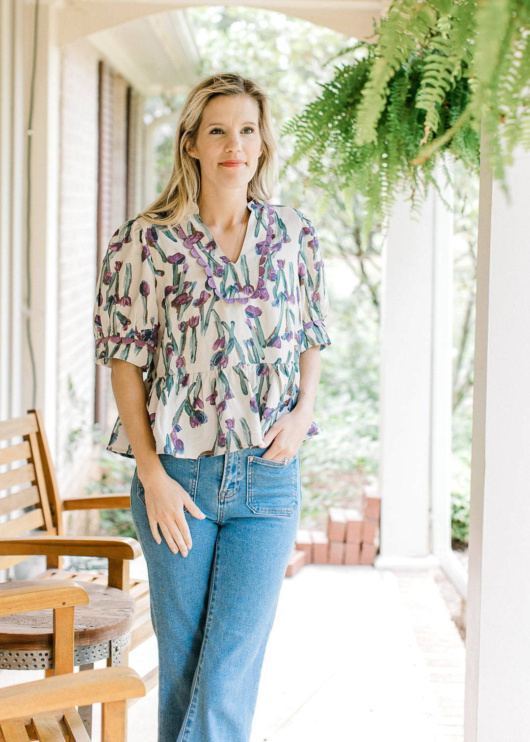 Model wearing jeans with a beige short sleeve babydoll top with lavender and blue floral. 