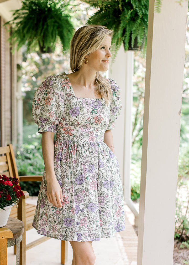Model wearing a short sleeve, off white above the knee dress with pink, purple and green floral. 
