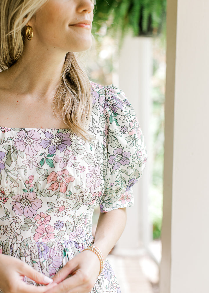 Model wearing a square neck, off white dress with colorful flowers and bubble short sleeve. 