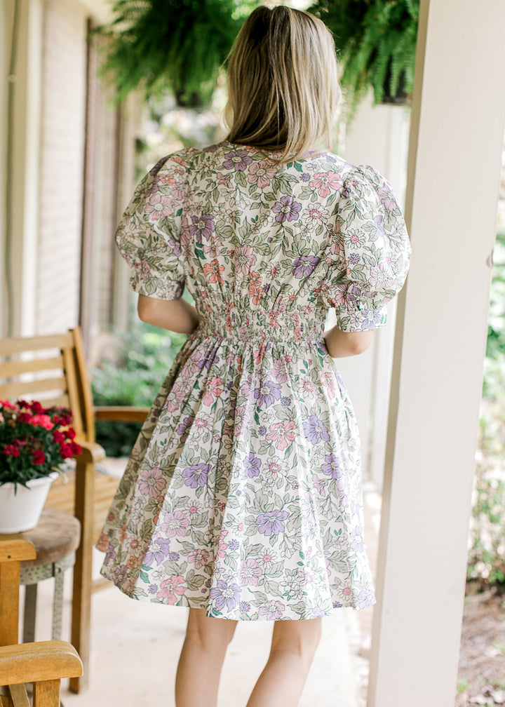 Back view of model wearing a short sleeve, off white dress with bright floral and a smocked waist.