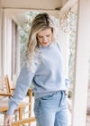 Model wearing a long sleeve dusty blue sweater with a crew neck and cream embroidered flowers. 