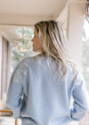 Back view of Model wearing a long sleeve dusty blue sweater with cream embroidered flowers. 