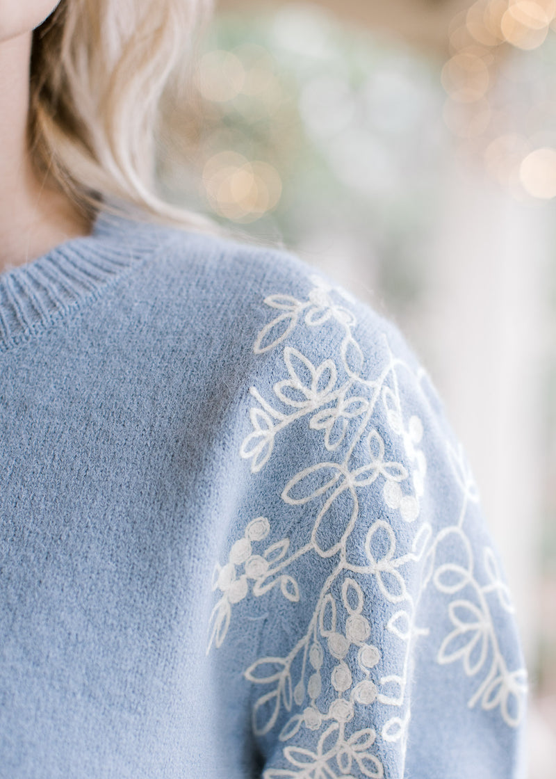 Detailed view of cream embroidery at the shoulder of a dusty blue long sleeve sweater. 