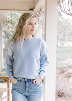 Model wearing jeans and a long sleeve dusty blue sweater with a cream embroidered flowers. 