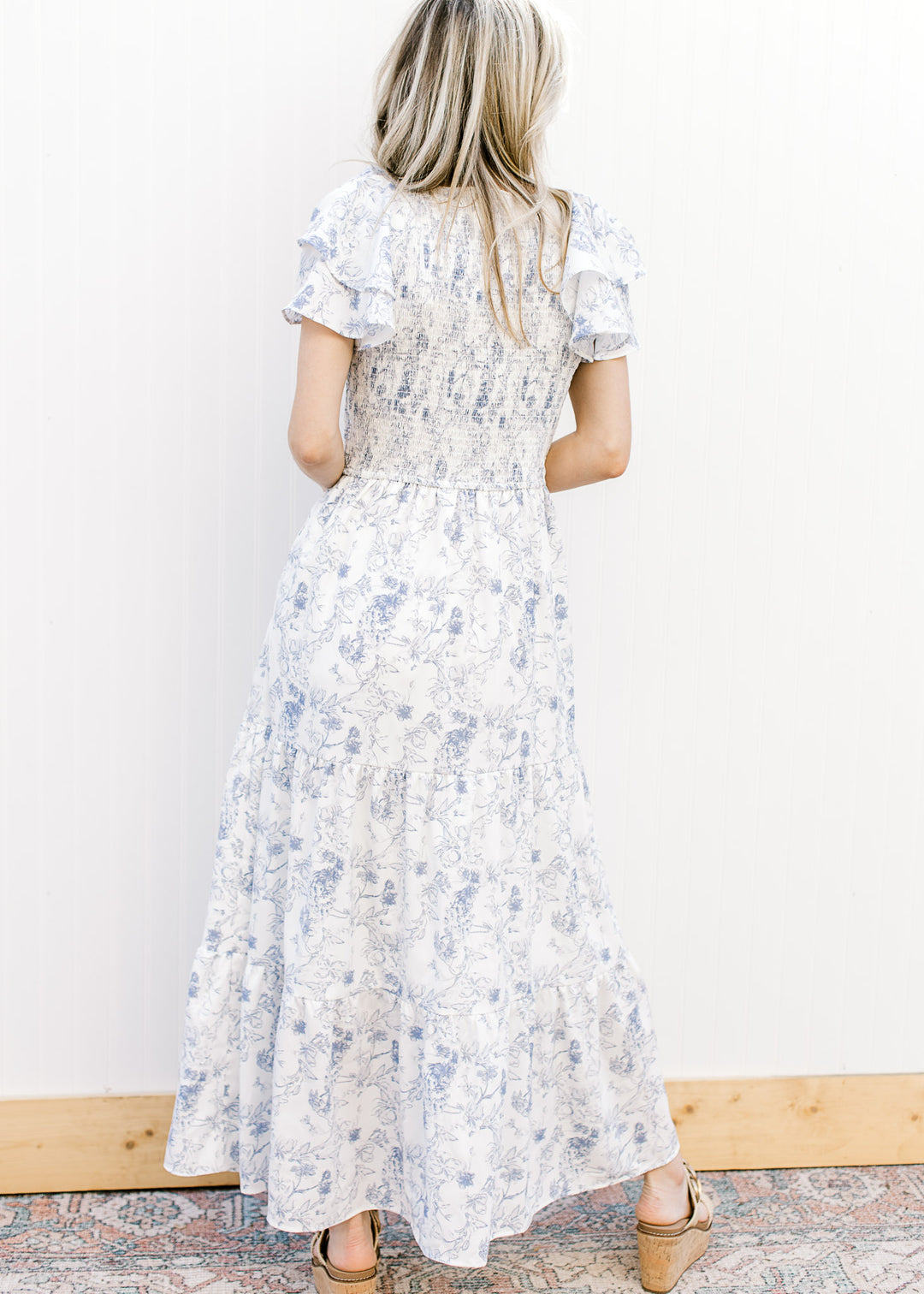 Back view of Model wearing a white maxi with blue floral, smocked bodice and ruffle short sleeves.