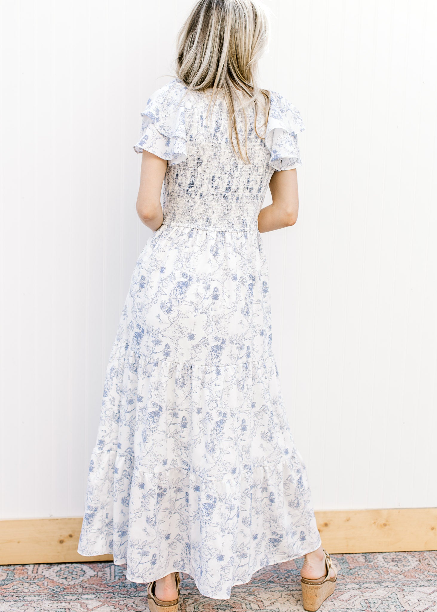 Back view of Model wearing a white maxi with blue floral, smocked bodice and ruffle short sleeves.